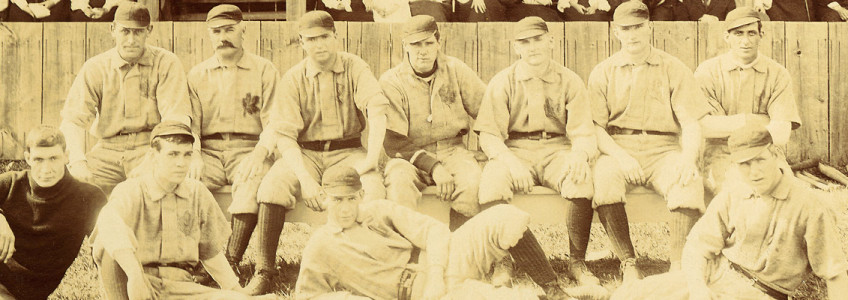 Sepia photo of a baseball team posing together on the baseball diamond. There are two rows of players wearing light coloured uniforms and high dark baseball socks and hats.
