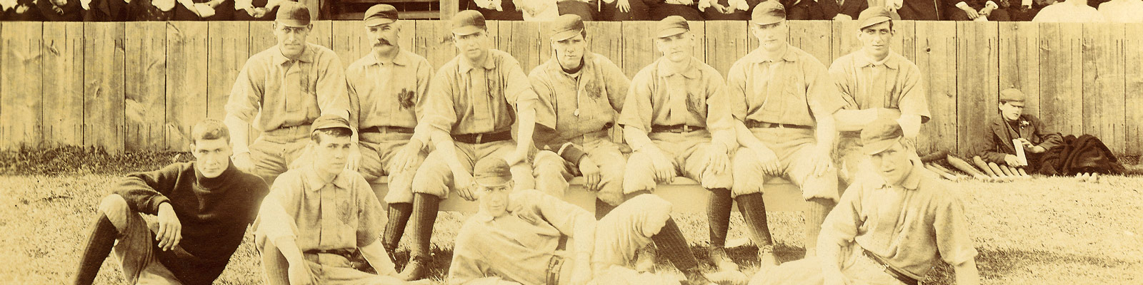 Sepia photo of a baseball team posing together on the baseball diamond. There are two rows of players wearing light coloured uniforms and high dark baseball socks and hats.