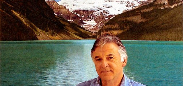 Photo of artist Ken Danby standing in front of a very blue lake. There are snow covered mountains in the background. Ken Danby has dark grey hair and is wearing a light blue collared shirt.