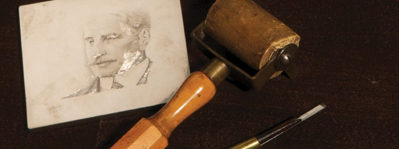 Engraving tools laid on a dark brown background. A black and gold pen or chisel tool is on the right, a rolling wooden mallet tool is in the centre, a white paper with a sketch of a man's face is on the far left.