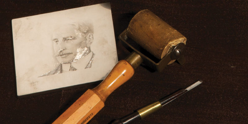 Engraving tools laid on a dark brown background. A black and gold pen or chisel tool is on the right, a rolling wooden mallet tool is in the centre, a white paper with a sketch of a man's face is on the far left.