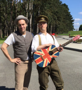 Murray Foster (left) and Mike Ford (right) standing in front of a forest with the vimy monument in the far background. Murray is standing with his hands on his hips, Mike is holding an acoustic guitar with the british flag design.