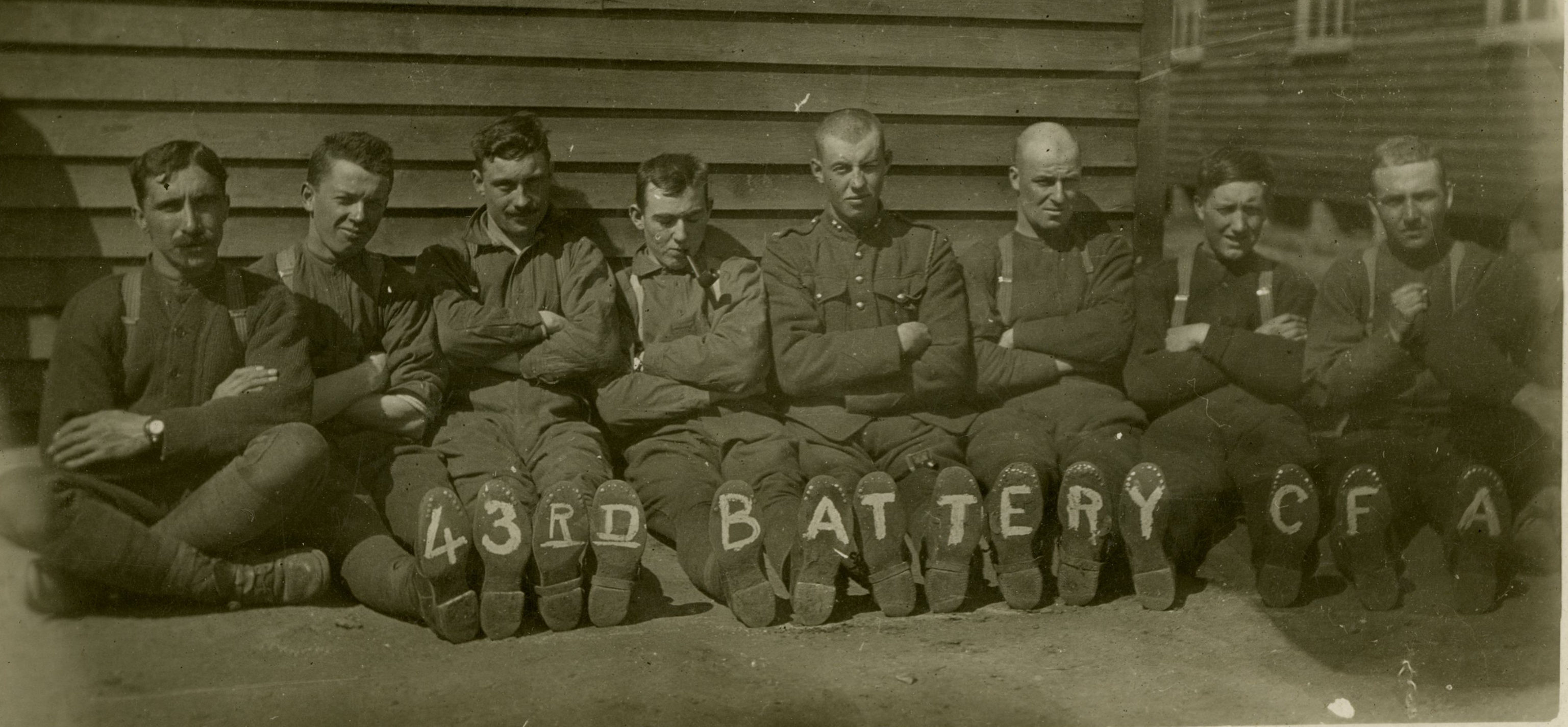 Sepia photo of seven soldiers sitting leaned up against a building. On the soles of their boots, each foot has a letter that fully reads 43rd Battery