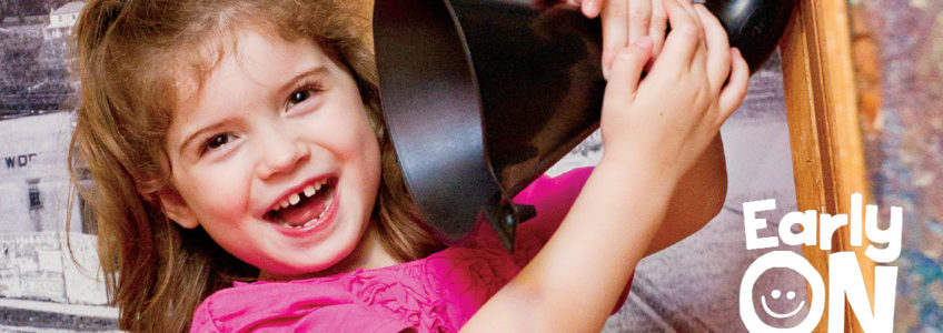 Photo of a young child smiling while playing inthe families gallery. She has brown hair and is wearing a hot pink shirt and is holding a black speaker to her ear. White text logo reads Early ON logo overlayed on the bottom right of the photo.