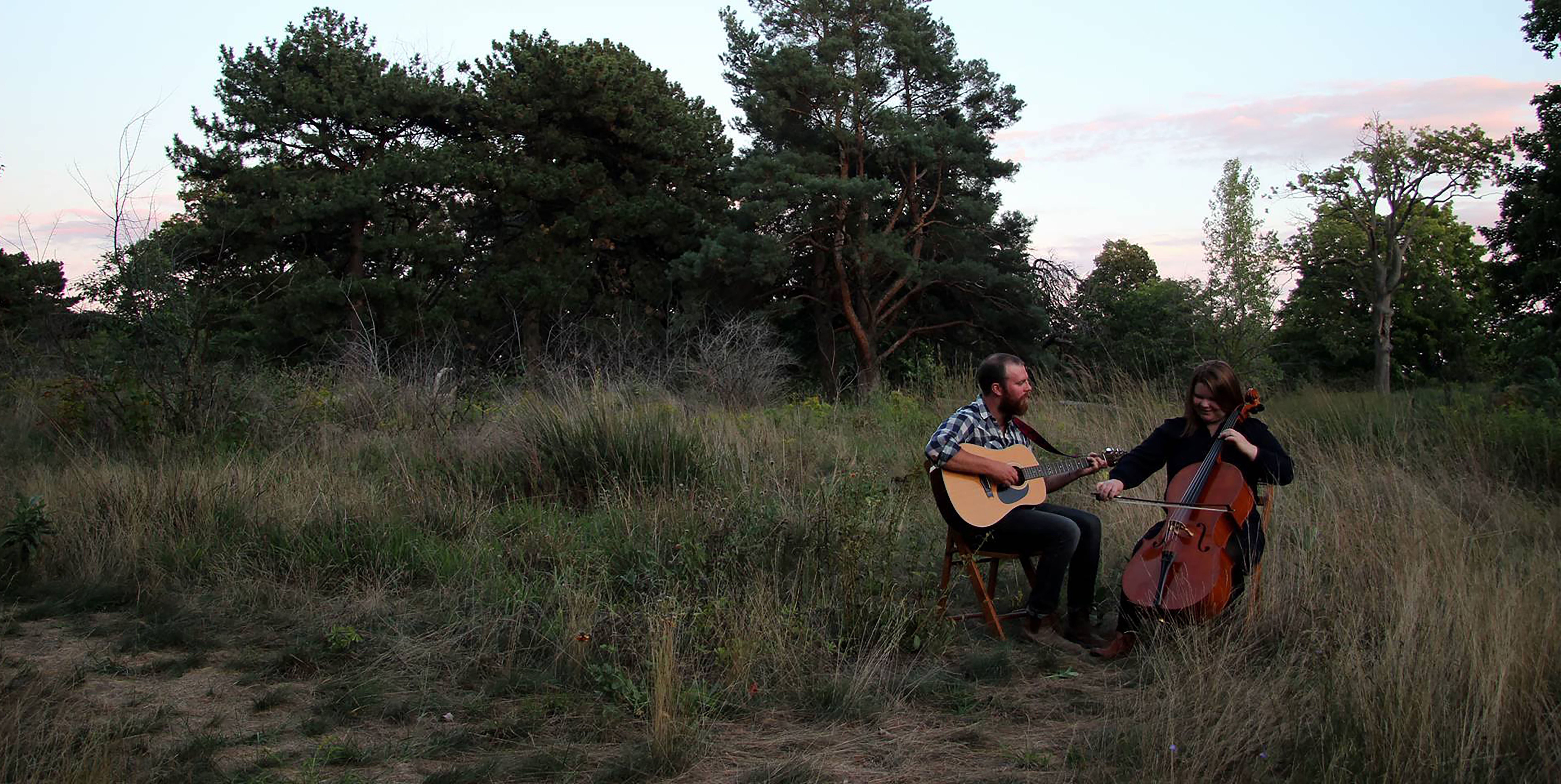 Photo of a man and a woman playing instruments while sitting in a field. The man on the left is holding a guitar and is wearing a grey collared shirt. The woman on the right is playing a cello and is wearing a black dress.