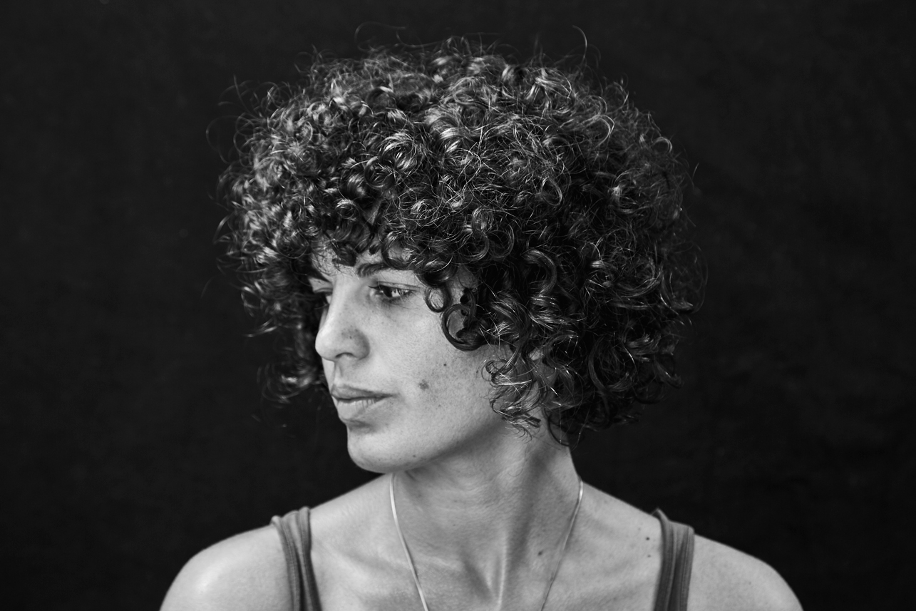 Black and white portrait photo of Ansley Simpson. Ansley has tight curly hair and is looking to the left. Ansley is sitting in front of a black background