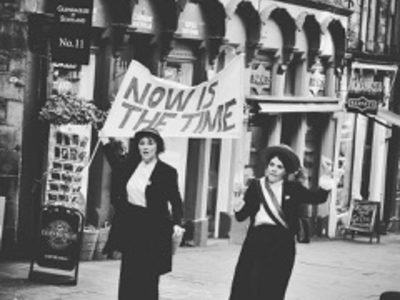 Black and white photo of two women dressed as suffragettes. They are wearing vintage style dress and skirts and are holding up their fists and are holding a sign that says now is the time.