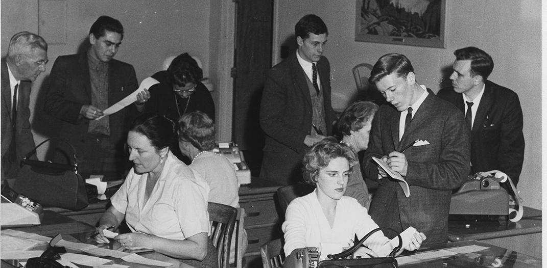 Guelph Mercury Newsroom circa 1960, two women sitting at desks typing, 5 men in suits standing and talking to each other.