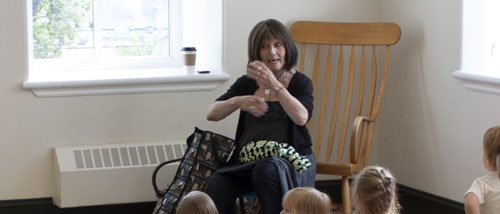 Jan sitting in a wooden rocking chair tellng a story to a group of young children.