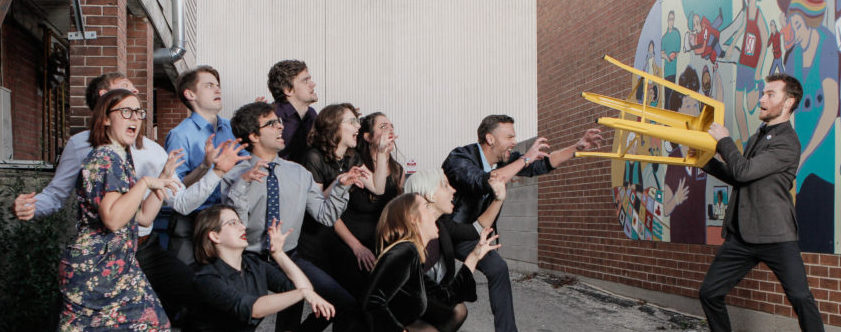 Photo of the Making Box Brigade. One man is holding up a yellow stool in defense against a large group of adults standing and crouching as if they were animals attacking him.