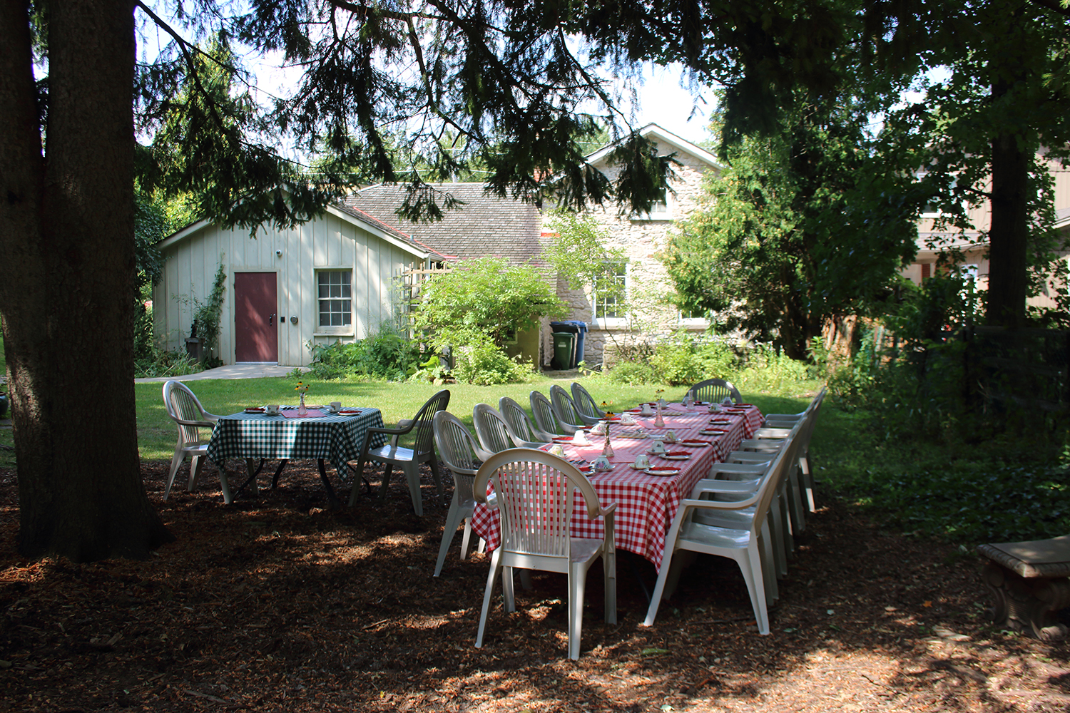 Two tables set up in the backyard gardens of McCrae House. Large pine trees frame the gardens.