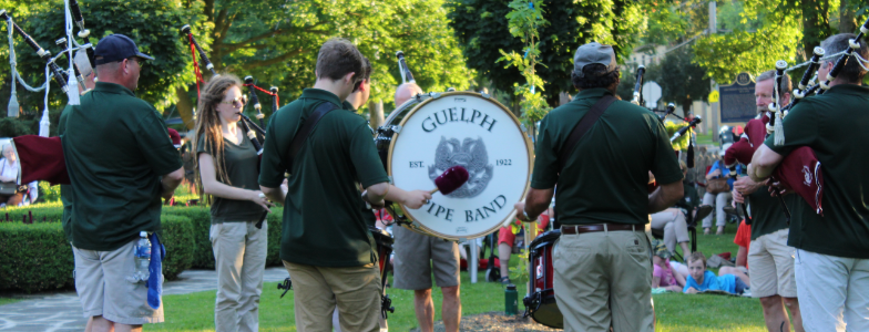 Photo of the Guelph Pipe Band standing in a circle while playing bagpipes. They are all wearing green shirts and khaki pants or shorts.