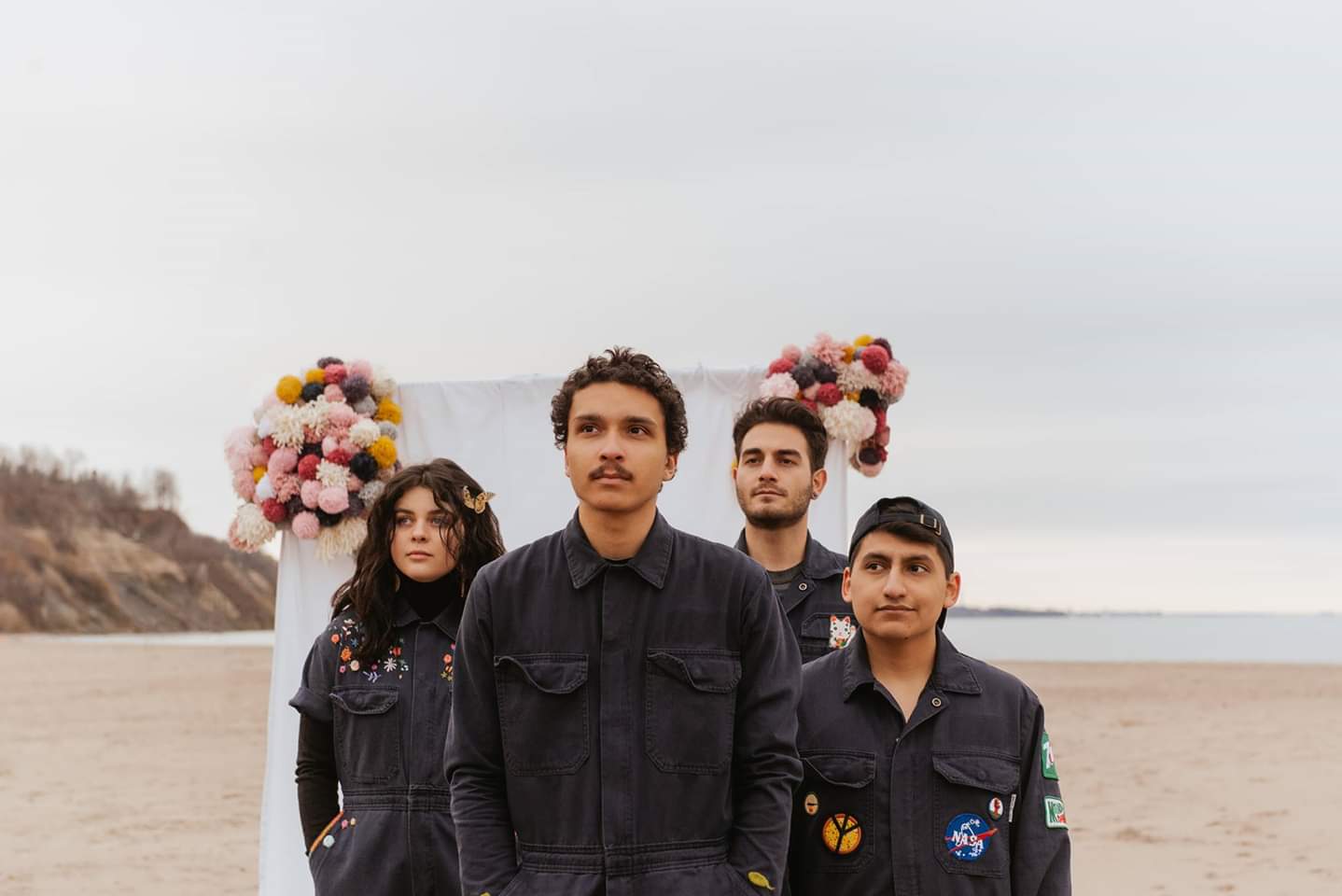Four young men and one woman standing together in black jumpsuits on a sandy beach. There is a white archway in the background with pink flowers on it.