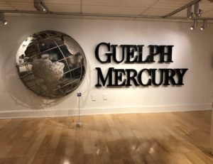 Guelph Mercury sign with globe originally from building. Large black metal letters "Guelph Mercury". The silver metal half of a globe sits to the left of the text and has an open cage design with flat textured metal pieces for the land masses.