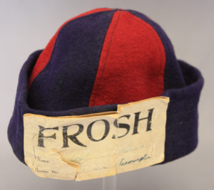Red and navy frosh beanie cap. The colours alternate around the centre a navy base. There is a yellowed label on the front that has FROSH written in large black writing.