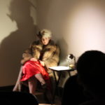 Close up of a storyteller dressed in a large fur coat sitting in front of visitors.
