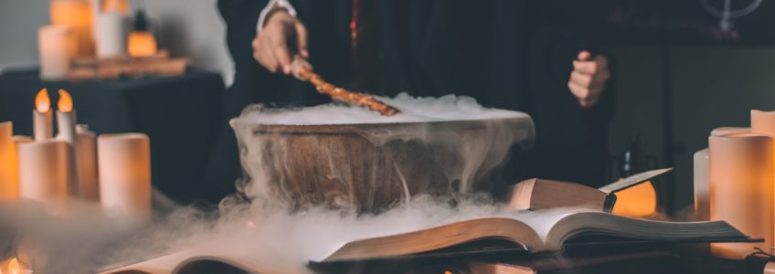 Stock photo of two people standing behind a cauldron wearing black Harry Potter robes. The cauldron is surrounded by white candles and a light fog.