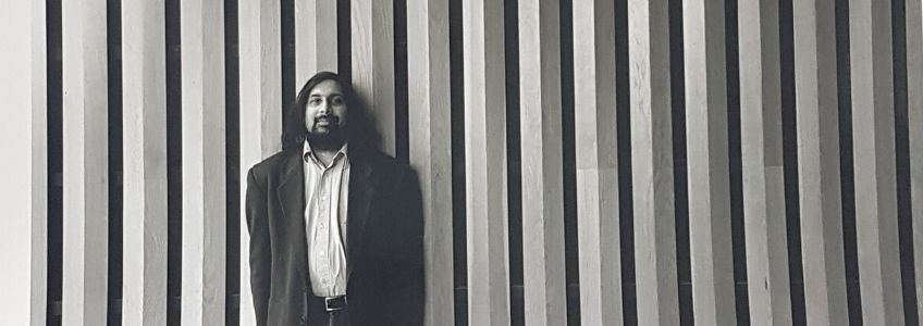 Ajay Heble standing in front of a paneled wall. He is wearing a dark suit jacket and a collared shirt.