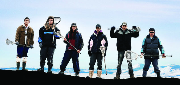 Group of six people holding lacrosse sticks standing facing forward. Snow covered mountains in the background.