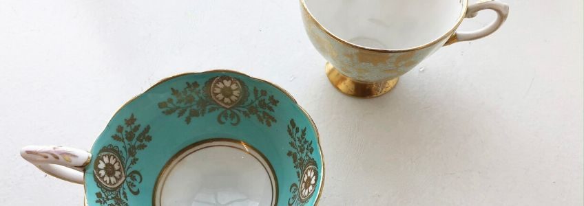 Photo of two china tea cups on a white background. Tea cup on the left is turquoise with gold floral details. The tea cup on the right is white with gold floral details.