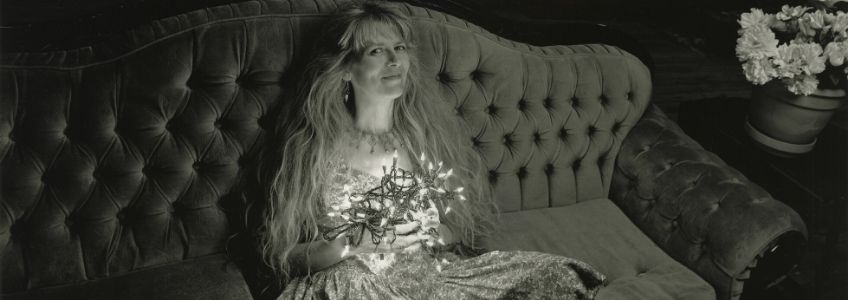 Black and white photo of Tannis sitting on a vintage looking couch. Tannis is holding a ball of white string lights.