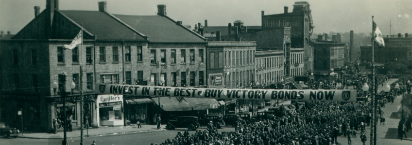 Black and white snapshot. Large group of people standing on Wyndham Street watching a parade. Buildings in background. Banner across the street "Invest in the Best Buy Victory Bonds Now". Band is leading the parade.