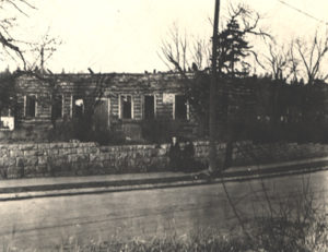 Black and white snapshot, with a white border, of a deteriorating log building with no roof, identified as the Priory, Guelph. A stone wall, utility pole and a road are in the foreground.