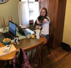 Sarah Ball's work from home setup. She is holding a tabby cat in her arms while sitting in front of her latop in the kitchen.