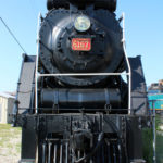 Photo of Locomotive 6167. The steam engine train is black with a red plaque under the headlight. On the red plaque, 6167 is written in gold.