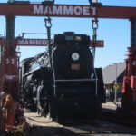 Photo of a black steam engine train, locomotive 6167, lifted up by a hydraulic lift system.