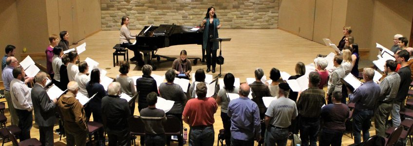 Photo of the Guelph Chamber Choir practicing. The large group is standing in a large semi-circle with their backs to the camera and are facing the conductor.