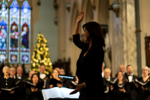 Charlene Pauls conducting in front of a large group of choir members. A christmas tree lit up with lights can be seen in the background.
