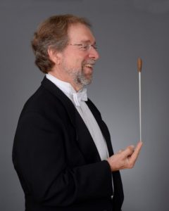 Conductor Gerald Neufeld wearing a black tuxedo. He is facing sideways and is balancing a conductor baton on his finger.