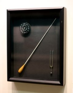Black shadow box display of a pitch pipe (left), conductor's baton (centre), and tuning fork (right)..