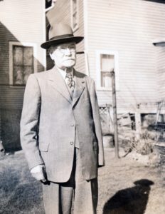 Portrait of Alfred “Fred” Hallett standing in a backyard. He is wearing a light coloured suit and brimed hat.