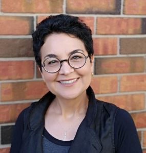 Image of Roxana Bahrami. She is wearing glasses and smiling at the camera. Her hair is short and she is standing in front of a brick wall.