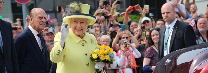 Queen Elizabeth II waving to a crowd of people in Berlin in 2015. She is dressed in yellow and holding yellow flowers.