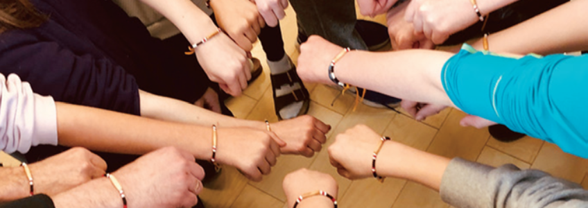 hairpipe bracelets, made by youth who took part in a workshop by Naomi Smith. Various arms are shown pointing in the center of a circle with their fists clenched. Bracelets are on their wrists.