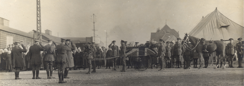 This is a glossy black and white photo. Rows of mourners (Nursing sisters, officers, soldiers) are seen in the background. In the foreground, 5 officers have their backs to the camera. 2 officers at the beginning of the procession are holding flowers and are pulling the cart holding McCrae's coffin. McCrae's coffin is covered with the Union Jack flag and Flowers. General Morrison (left) and General Aime (right) are in the front row of mourners, seen between Bonfire and the coffin. General Arthur Currie is standing at attention in the line of officers. He is right at Bonfire's nose. The nursing sisters are to the left.