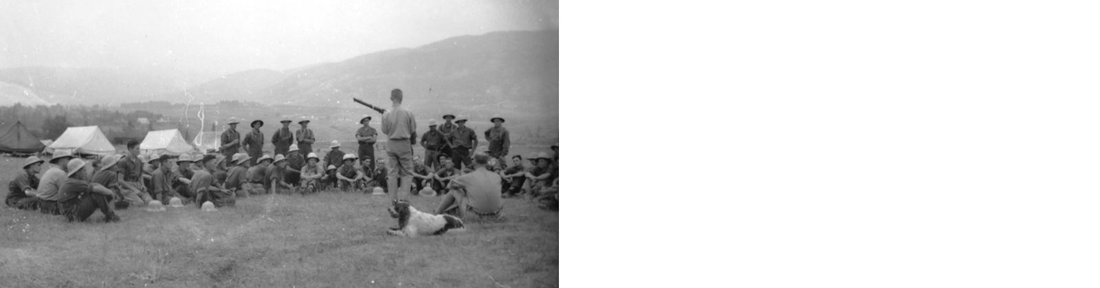 a group of men sit in a field watching a field lecture. A man is demonstrating something infront of them. A dog is in the foreground.