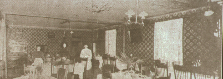 Digital image of the interior of the American Hotel. The images shows the dining room in the hotel with two ladies standing at the back. The room has wood floors and is set with tables and chairs. The tables have light colured table cloths on them and flowers in the centre of each table. The tables are also set with utensils. Hanging from the ceiling is a gas two branch lamp. The walls have patterned wallpaper.