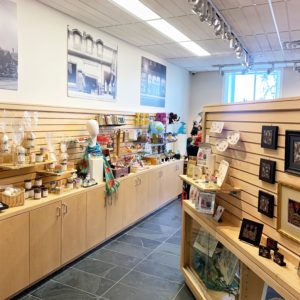 The inside of the Hilltop Shop at the Civic Museum. Merchandise is displayed on light wood shelfing. Various items like paintings, gifts, scarves, and children's toys are available. Above the shelves hang large pictures of old Downtown Guelph. Light is coming in from the back window.