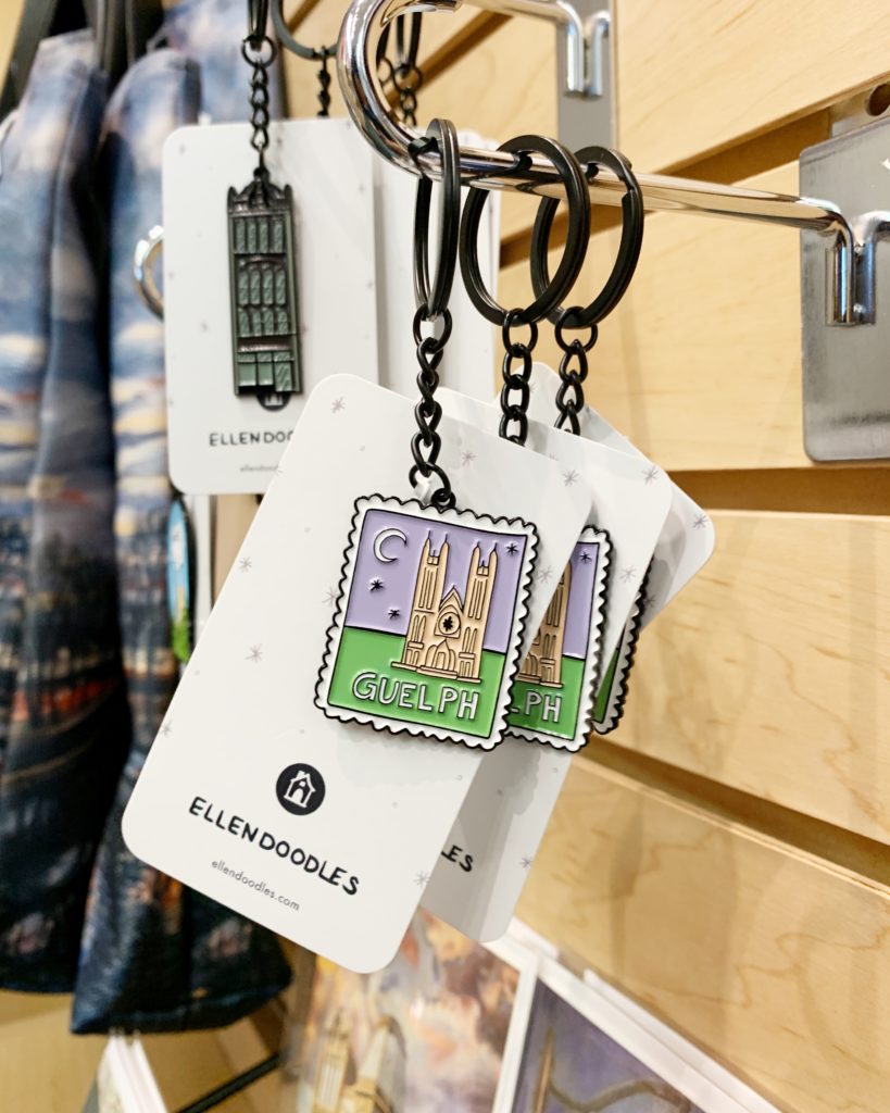 Keychains hanging from silver hooks in the Hilltop Shop. The front keychain is a design of the Basilica of Our Lady, and behind it hangs a design of the Petrie Building. 