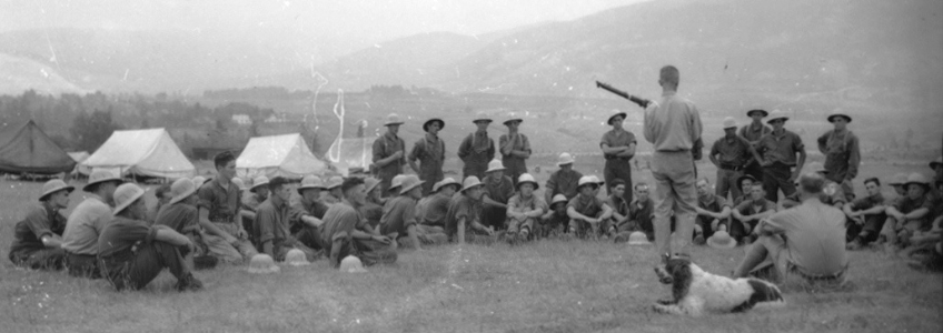 Group of soldiers laying on a field, they are watching one man give a demonstration/lecture on training.