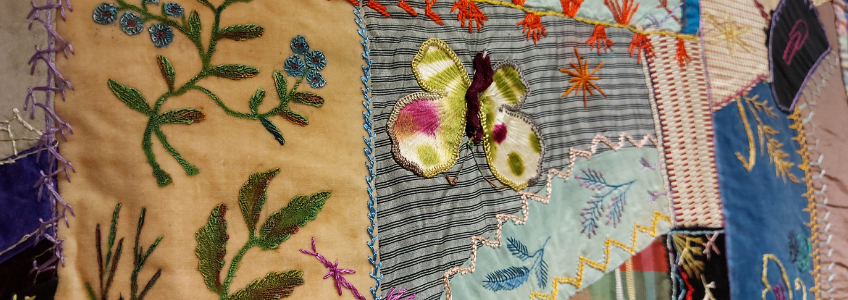 Detailed view of Crazy Quilt. Different panels of fabric are shown, one which includes an embroidered butterfly and floral designs.