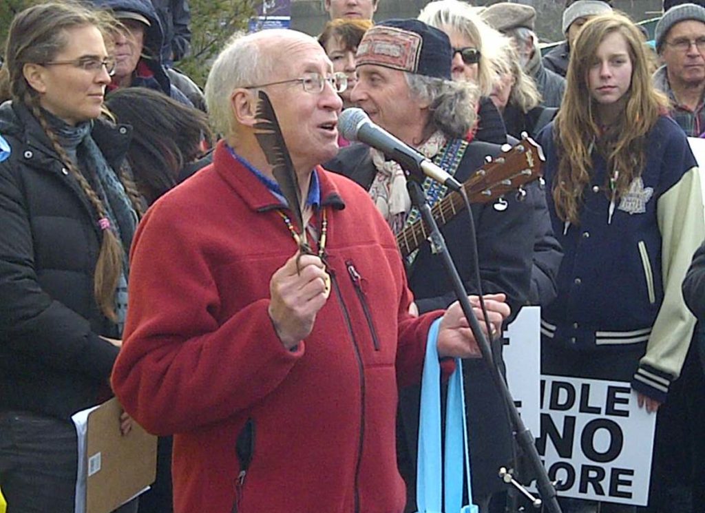 Bruce Weaver, wearing a red jacket and holding a long white feather, stands speaking into a microphone. Several individuals including James Gordon, stand in a group behind the speaker, listening to the conversation. In the background, a large glass windowed building can be seen.