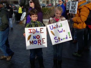 Two children stand amongst a group of people. The children are carrying signage which reads "Idle No More" and "United We Stand" In the background several other signs reading "Idle No More" can be seen, held by some members of the group.