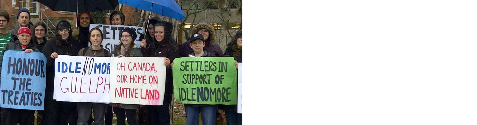 A group of people wearing winter clothing stand together on a ice and snow covered grassy area located in front of Zavitz Hall at the University of Guelph. Several signs are held by the smiling participants and read "Honour The Treaties", "Idle No More Guelph", "Oh Canada, Our Home On Native Land", and "Settlers In Support of IdleNoMore".