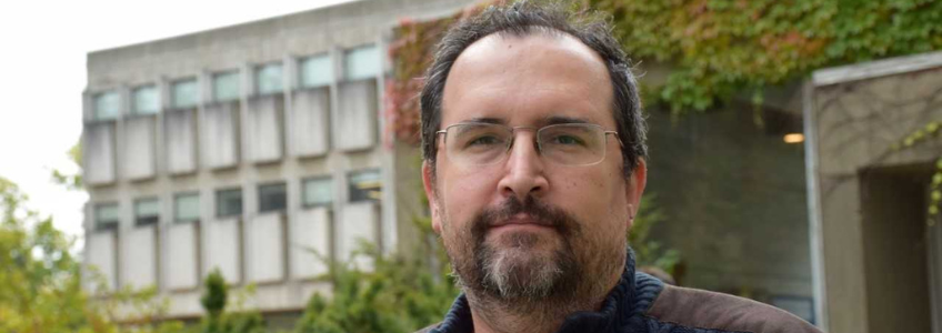 Evren Altinkas outside a university building. Her is wearing a blue cardigan with large buttons. He has short brown hair and a goatee. Behind him are buildings and a tree.