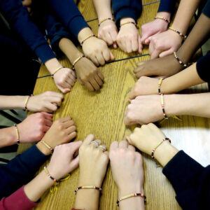Image of human hands in a circle and on a wooden table. Each wrist has a beaded bracelet. 